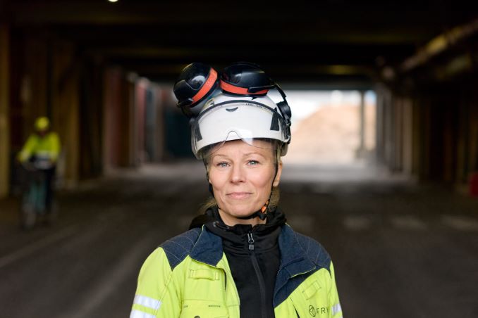 En kvinna som har på sig skyddshjälm, skyddsglasögon och en varseljacka med AFRY-logga tittar in i kameran. I bakgrunden syns en tunnelkonstruktion.